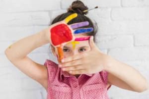 portrait of girl looking through her painted hands standing against white brick wall