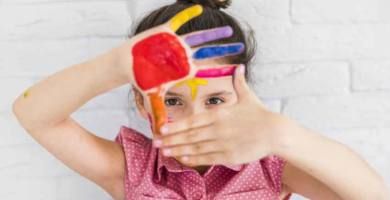 portrait of girl looking through her painted hands standing against white brick wall